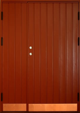 Pardörr i rödbrunt trä med stående panel och enkel design. Dörrarna har metallbeslag vid låset och förstärkning i form av kopparplåtar längst ned. Svarta gångjärn syns på båda sidor.