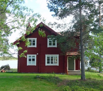En röd timrad tvåvåningsvilla med vita fönster och grönt dörrparti, belägen i en naturnära miljö med gräsmatta och höga träd. I bakgrunden syns en sjö som speglar himlen. Huset har spröjsade fönster i klassisk stil och ett rött tegeltak.