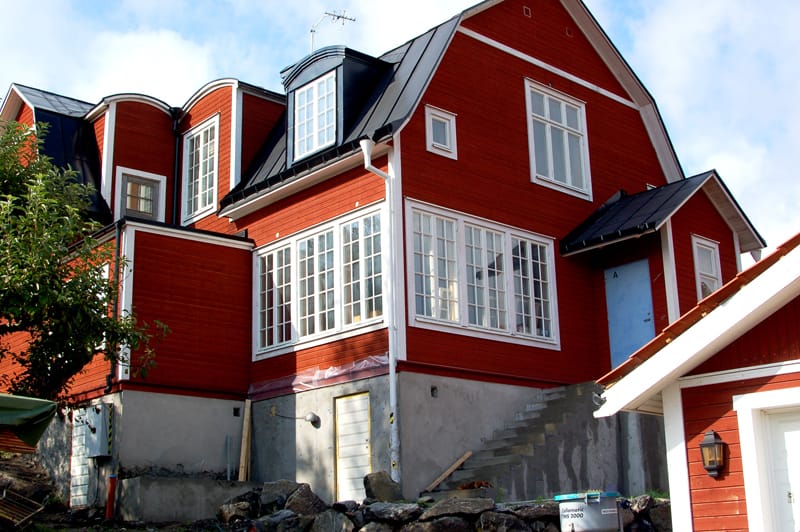Ett stort rött trähus med vita foder runt spröjsade fönster och svart plåttak. Huset har flera burspråk och takkupor med fönster samt en glasveranda som löper längs ena sidan. Sockeln är i betong och det pågår byggnation vid grunden, med synliga stenar, verktyg och en ofärdig trappa upp mot entrén. I förgrunden syns delar av ett mindre rött uthus med vitmålad takkantslist.