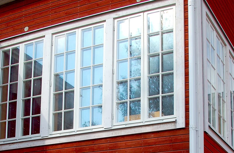 Närbild av en glasveranda på ett rött trähus med vitmålade spröjsade fönster. Fönstren är stora, går nästan från golv till tak och består av flera smala glasrutor i klassisk stil. De vita foderna kontrasterar mot den röda träpanelen, och glaset speglar träd och himmel utanför.