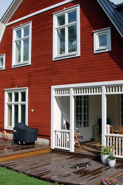 Rött trähus med vita detaljer och flera höga spröjsade fönster. På övervåningen sitter två höga tvådelade fönster samt ett litet fyrkantigt fönster. På nedervåningen finns ett större fyrdelat fönster samt en altan med tak och vita räcken. På altanen står stolar och bord, en grill är placerad på trädäcket och en skateboard ligger på marken.