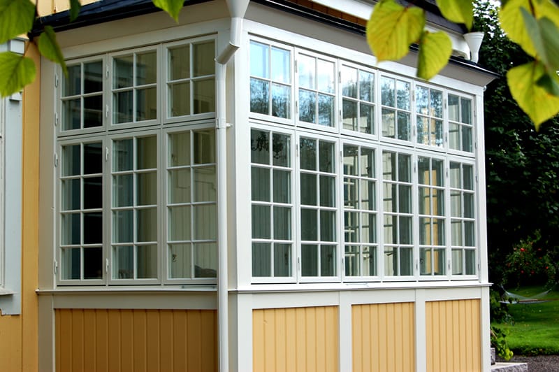 En glasveranda på ett gult trähus med vitmålade spröjsade fönster som täcker två väggar. Verandan har en vit nedre del och takränna, och resten av huset syns med gul panel. Utanför finns grönska och träd, vilket skapar en lantlig och idyllisk känsla.