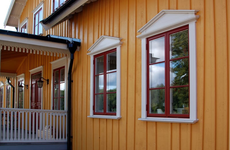 Närbild på en gulmålad trävilla med röda fönsterbågar och vita omfattningar. Ovanför fönstren sitter dekorativa vita överstycken med trekantiga gavelmotiv. Till vänster syns en vitmålad veranda med snickarglädje och svarta vägglampor.