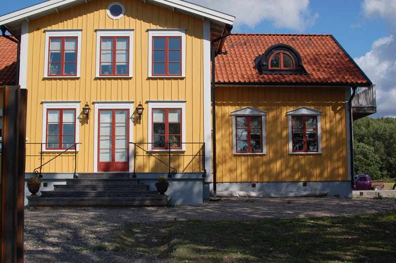 Tvåvåningshus med gul träfasad och rött tegeltak. Huset har flera rödmålade fönster med vita omfattningar, samt en vit dubbeldörr med glasrutor i mitten. Till höger syns ett svart rundbågefönster på övervåningen. Framför entrén finns en trappa med järnräcken och två blomkrukor.