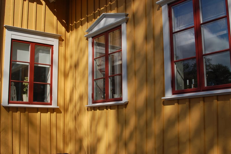 Närbild av gulmålad träfasad med stående panel och tre rödmålade fönster med vita omfattningar. Det mittersta fönstret har ett vitt triangulärt överstycke medan de andra har raka överstycken. Solen kastar skuggor från träd över fasaden.