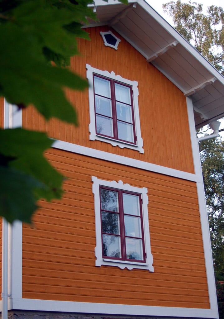 En orange träfasad med två rödmålade fönster, ett på övervåningen och ett på nedervåningen. Fönstren är indelade i fyra rutor med spröjs och har dekorativa, vita snickarglädjeomfattningar. Under taknocken sitter dessutom ett litet sexkantigt fönster med vit inramning. Gröna löv syns delvis i förgrunden till vänster.