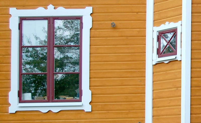 Två fönster på en fasad med orange träpanel. Till vänster ett större rektangulärt fönster med rödmålad båge och vit profilerad dekorlist, indelat i fyra rutor med spröjs. Till höger ett mindre fyrkantigt fönster med samma färgsättning, indelat i fyra rutor med krysspröjs. Båda fönstren har vita dekorativa foder.
