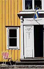 Gul träfasad med vit dekorerad ytterdörr, svensk flagga ovanför och vit stol med röd- och vitrandig dyna vid sidan om.