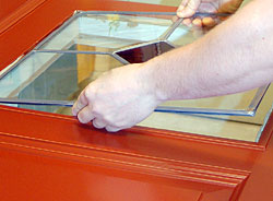 Närbild av röd spegeldörr där en person monterar en glasruta med rombformad spröjskassett i den övre spegeln.