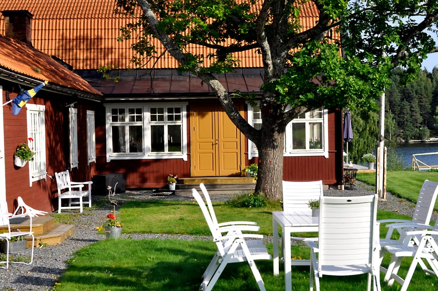 Rött trähus med gul pardörr och vitmålade spröjsade fönster, omgivet av gräsmatta, träd och vita trädgårdsmöbler nära vatten.