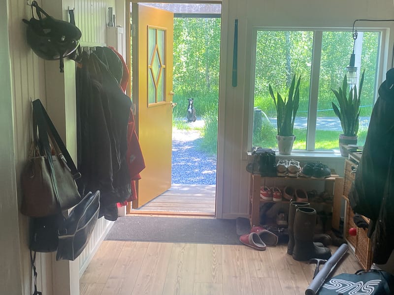 Inomhusvy mot öppen gul ytterdörr med diamantformat glas i dörrbladet, med fönster i ljus karm som släpper in dagsljus.
