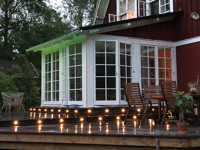 Vit glasveranda med spröjsade fönsterdörrar och fönsterpartier mot en altan, där kvällsljus och lampor speglas i glasen och i det blöta trädäcket.