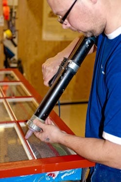 En snickare står i en verkstad och arbetar koncentrerat med en svart fogspruta. Han applicerar fogmassa längs kanten på ett rött fönster- eller dörrparti med glas, som ligger på ett arbetsbord. Personen bär arbetskläder och glasögon, och miljön är en hantverksverkstad med fokus på noggrant och detaljerat montagearbete.