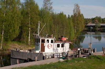En liten arbetsbåt ligger förtöjd vid en träbrygga en grönskande kväll. Båten är vit med detaljer i rött och svart och omges av lugnt vatten som speglar träd och himmel. Runt sjön står täta björkar och lövträd i vår- eller försommargrönt. Stämningen är stillsam och lantlig.