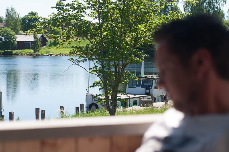 En suddig person i förgrunden sitter i skugga medan blicken öppnar sig mot ett somrigt svenskt landskap. I bakgrunden syns en stilla sjö med en förtöjd båt, gröna träd och röda hus på andra sidan vattnet. Solen lyser starkt och scenen förmedlar lugn, sommarvärme och en känsla av ro vid vattnet.
