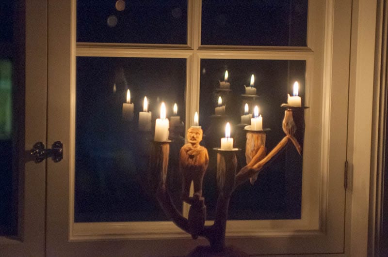 Ett fönster i mörker med tända stearinljus i en traditionell träljusstake. Ljusens lågor speglar sig i fönsterglaset och skapar ett varmt sken mot den mörka februarinatten utanför.