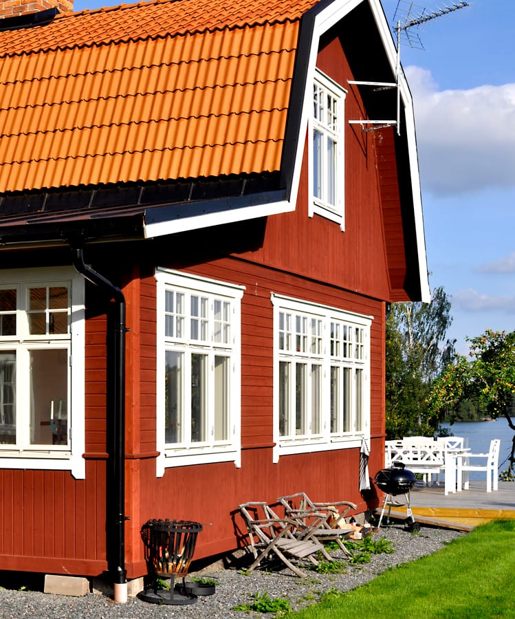 Rött trähus med vitmålade spröjsade fönster och gavelfönster, täckt av tegeltak, beläget i grön trädgård med altan och utemöbler.