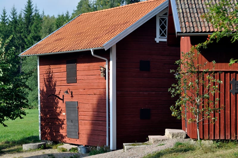 Rödmålad träbyggnad med tegeltak och enkla fönsterluckor, belägen i lantlig miljö med grönska och träd runt omkring.